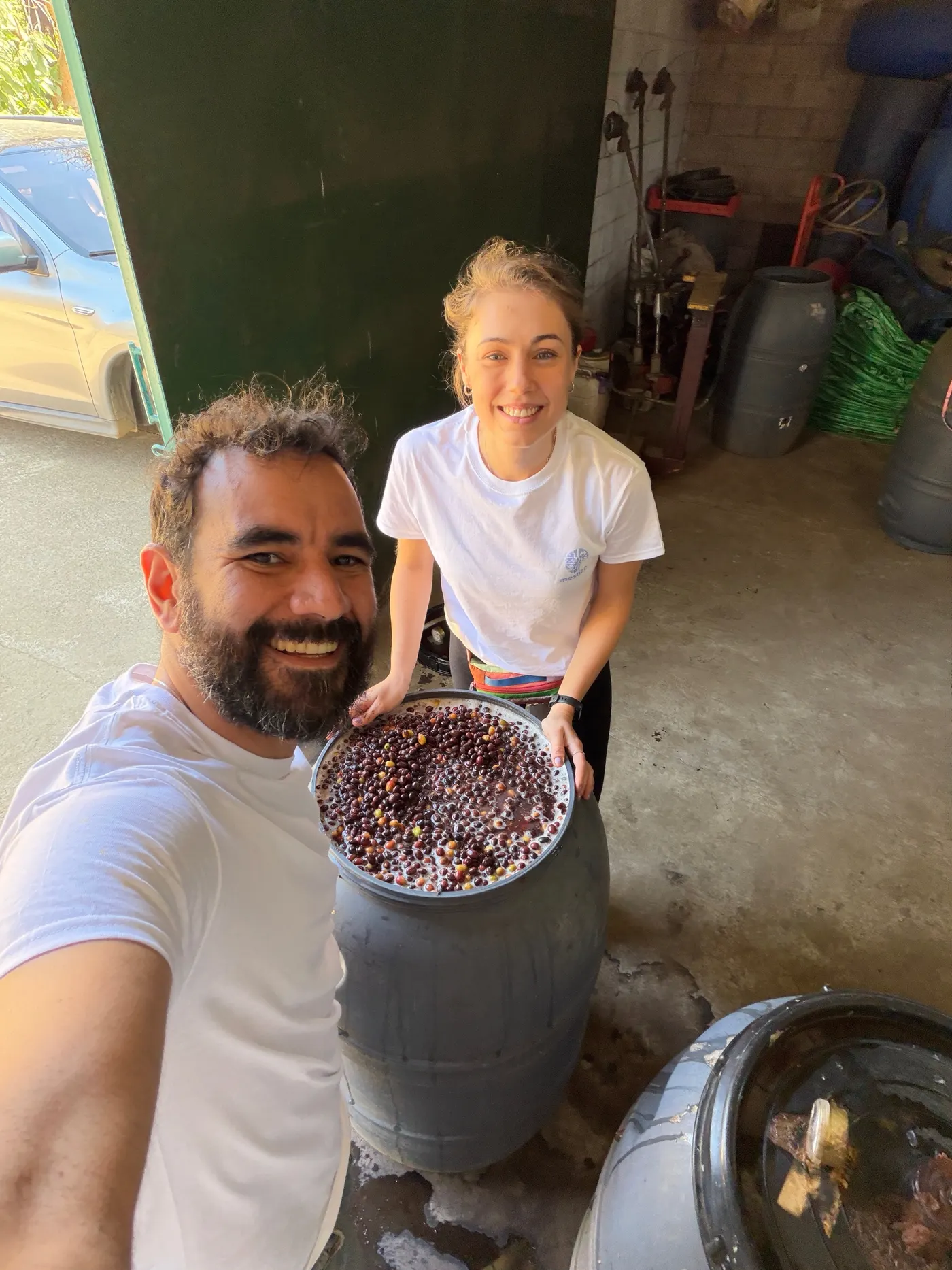 Miguel and Joanna standing together beside an open drum of coffee cherries, smiling at the camera in an open sided shed with a concrete floor, tools and more drums behind them.