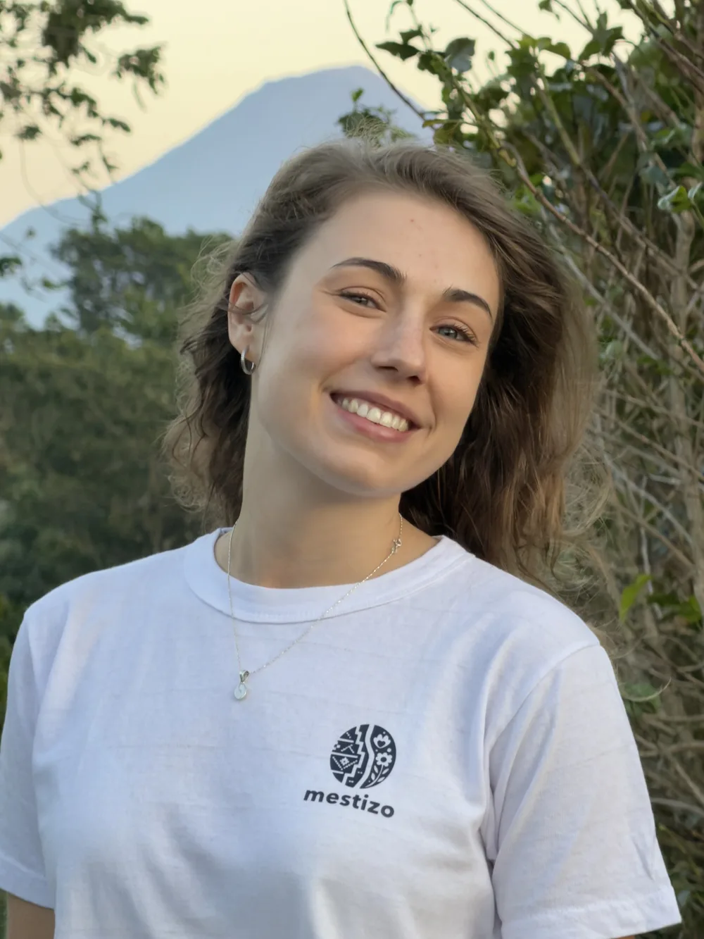 Joanna outdoors at golden hour in a white Mestizo t-shirt, the same volcano behind her.